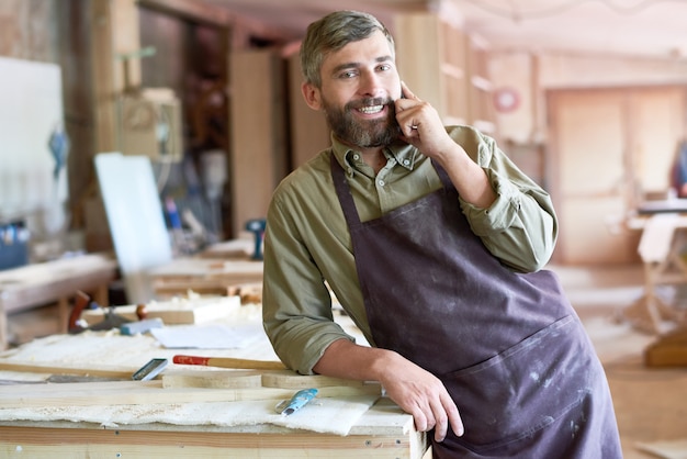 Carpentiere barbuto che parla da Smartphone in officina moderna