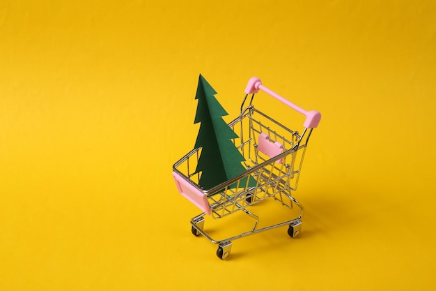 Carrello del supermercato con carta tagliata albero di Natale su sfondo giallo Shopping di Natale