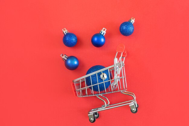 Carrello del supermercato e palle di Natale blu su sfondo rosso. Minimalismo, copia spazio, piatto laici.