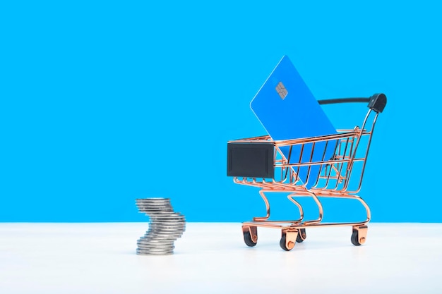 Carrello della spesa giocattolo con una carta di credito e monete su un concetto di shopping isolato su sfondo blu