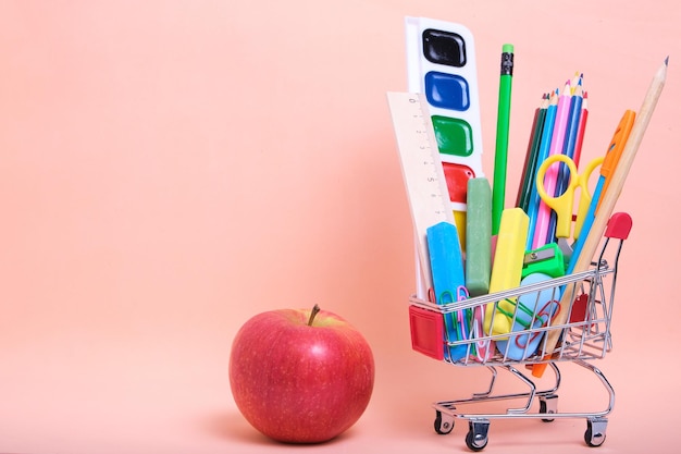 Carrello della spesa materiale scolastico completo e forniture per ufficio e sfondo rosa mela rossa