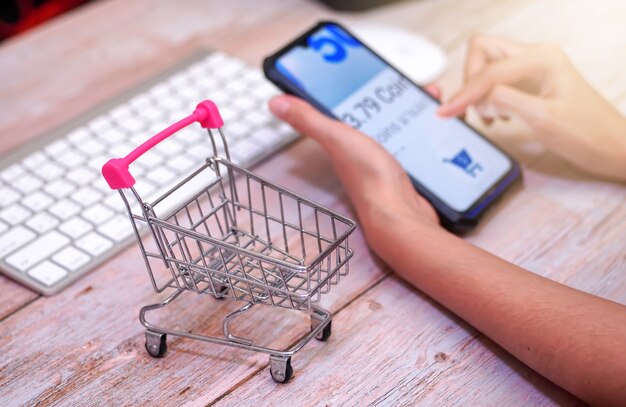 carrello della spesa su un tavolo di legno Dietro una donna che utilizza uno smartphone sfocato shopping online