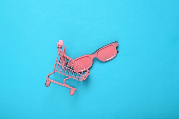 Carrello mini supermercato rosa con occhiali da sole rosa su sfondo blu Vista dall'alto Piatto Concept art Minimalismo