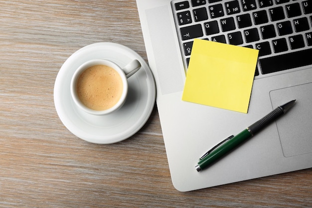 Carta adesiva gialla vuota su penna da tastiera per laptop e tazza da caffè sullo sfondo della scrivania