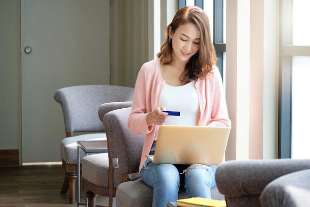 Carta di credito della tenuta della giovane donna per la compera online mentre usando i computer portatili nell'umore sorridente.