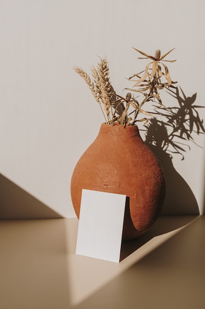 Carta di foglio di carta bianca con spazio di copia e vaso di terracotta con bouquet di grano secco con ombra di luce solare su bianco