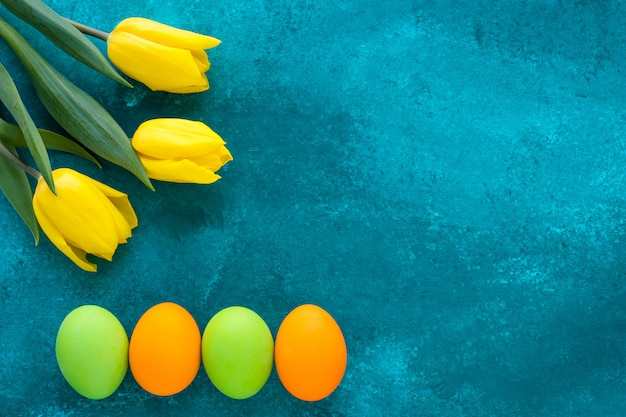 Carta regalo pasquale con uova dipinte luminose e tulipani gialli su sfondo turchese scuro grunge. Cornice di Pasqua festiva con spazio di copia.