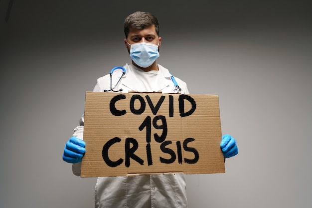 Cartello "crisi Covid-19" nelle mani del medico uomo manifestante su sfondo grigio.