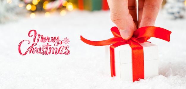 Cartolina d'auguri di buon Natale con una mano di uomo che tiene una confezione regalo con nastro rosso sulla neve e luci sfocate