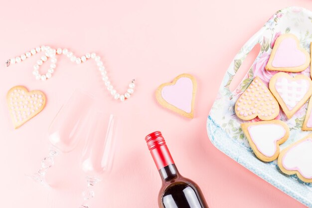 Cartolina d'auguri di buon San Valentino con biscotti del cuore, bicchieri di vino e vino su sfondo rosa pastello.