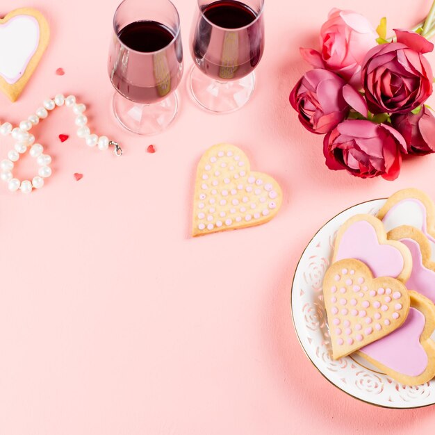 Cartolina d'auguri di buon San Valentino con biscotti del cuore, bicchieri di vino e vino su sfondo rosa pastello.