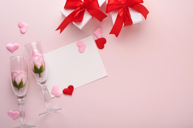 Cartolina d'auguri di San Valentino con bottiglia di champagne, confezione regalo, nastro rosso e busta con mockup di note vuote all'interno su sfondo rosa. Vista dall'alto con spazio per i saluti. Biglietto di auguri con spazio per la copia