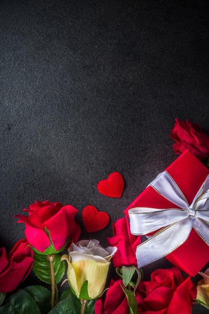 Cartolina d'auguri di San Valentino con bouquet di fiori di rosa rossa, vino e confezione regalo sul tavolo nero. Vista dall'alto con spazio per i saluti