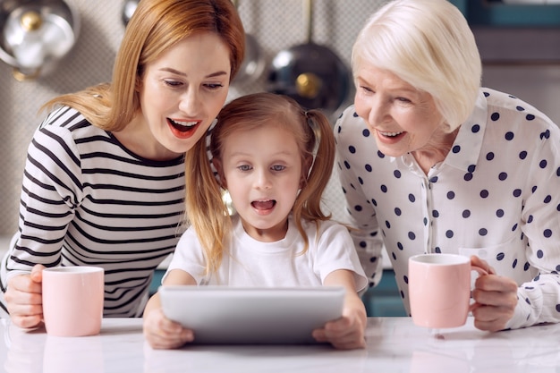 Cartone animato emozionante. Piacevole bambina che guarda un cartone animato su tablet insieme a sua nonna e madre che bevono caffè da tazze rosa mentre guarda divertito da colpi di scena