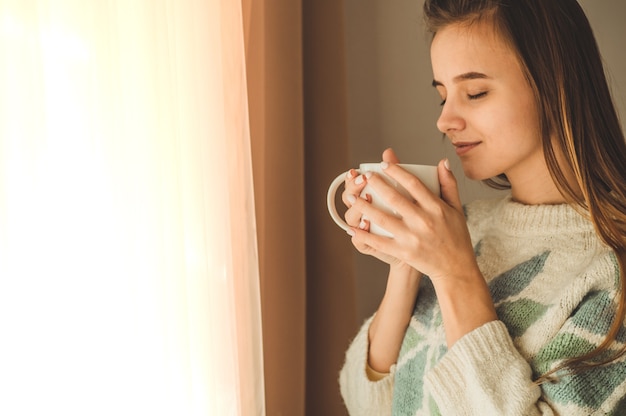 Casa accogliente. Donna con una tazza di bevanda calda vicino alla finestra. Guardando la finestra e bevi il tè. Buongiorno con il tè. Donna abbastanza giovane che si distende. Felice concetto.