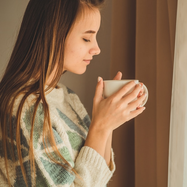 Casa accogliente. Donna con una tazza di bevanda calda vicino alla finestra. Guardando la finestra e bevi il tè. Buongiorno con il tè. Donna abbastanza giovane che si distende. Felice concetto.