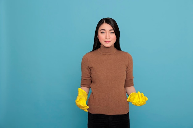 Casalinga asiatica sorridente felice dalle faccende domestiche che indossa guanti gialli in piedi su uno sfondo blu, concetto di casa di pulizia, allegro soddisfatto e felice con uno stato d'animo positivo