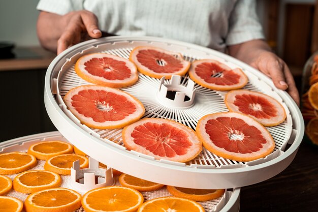 Casalinga contemporanea in piedi accanto al tavolo della cucina e mettendo il vassoio dell'essiccatore di frutta con pompelmo a fette su quello con fette d'arancia