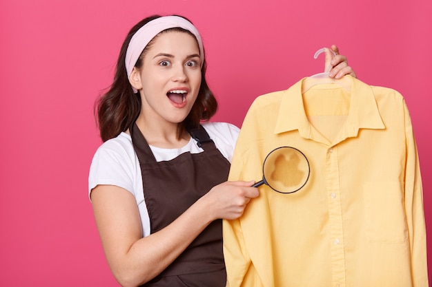 Casalinga dall'aspetto piacevole con sguardo stupito, indossa una fascia per capelli bianca, una maglietta e un grembiule marrone, la donna mostra una grande macchia con lente d'ingrandimento, ha bisogno di rimuovere le impurità.