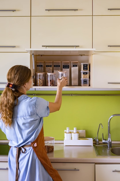 Casalinga femminile che mette barattoli di vetro pieni di cibo sano, noci, cereali, cereali sullo scaffale dell'armadio