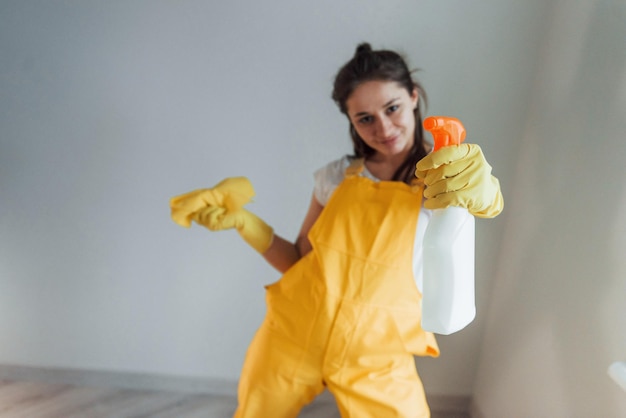 Casalinga in uniforme gialla in piedi con spray detergente per finestre all'interno Concezione di ristrutturazione della casa