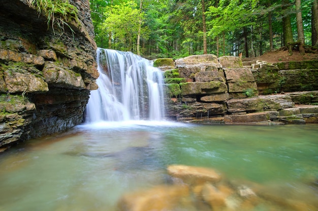 Cascata sul fiume di montagna con acqua schiumosa bianca che cade dalla formazione rocciosa nella foresta estiva.