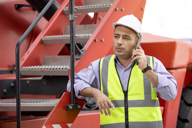 Casco dell'operaio in piedi e parlando al telefono cellulare al cantiere di spedizione di container di carico logistico