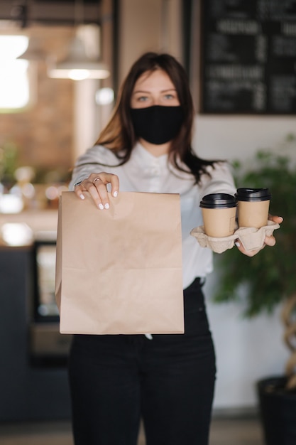 Cassiere in maschera facciale tenendo il pacchetto con il cibo e due tazze di caffè concetto pagamenti senza contatto