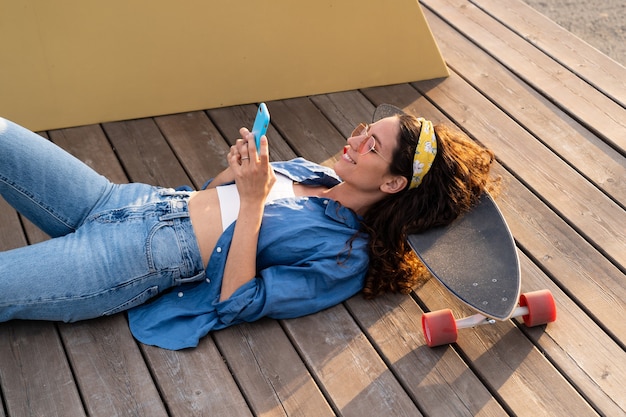 Casual giovane ragazza hipster in occhiali da sole che sms in smartphone all'aperto sdraiato rilassato in longboard