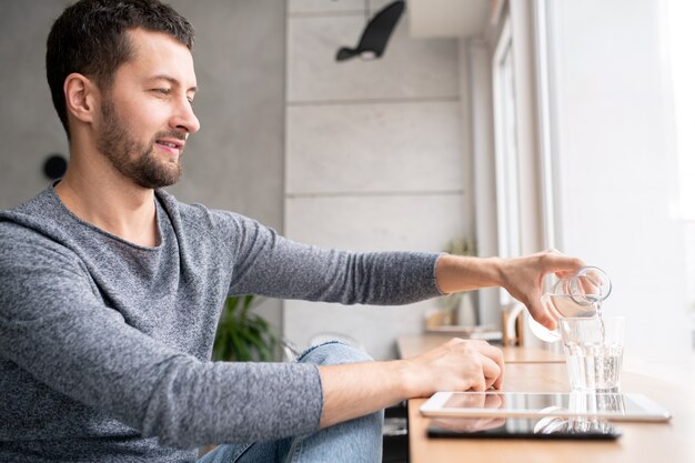 Casual giovane uomo seduto dalla finestra nella caffetteria e versando acqua dalla bottiglia in vetro durante una breve pausa