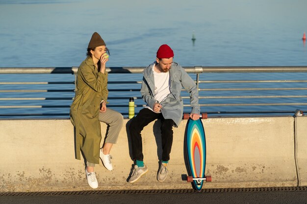 Casual hipsters ragazzo e ragazza alla data insieme bevono tè seduti sul ponte sul fiume al tramonto sun