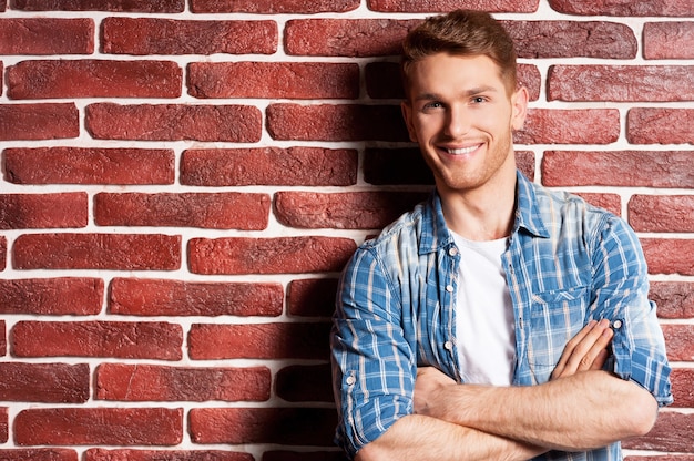 Casualmente bello. Bel giovane che tiene le braccia incrociate e guarda la telecamera mentre sta in piedi contro un muro di mattoni