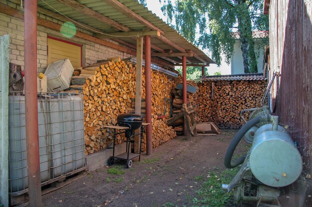 Catasta di legna sul muro della casa con una griglia e un serbatoio dell'acqua. La legna da ardere è coperta da un tetto. La scatola e i contenitori mentono. Appendere il bordo delle ruote. Dietro la recinzione cresce una spessa betulla.