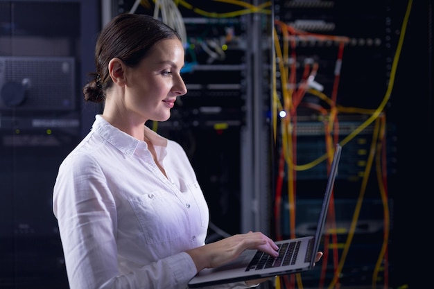 caucasian female engineer using laptop in computer server room. database server management and maintenance concept