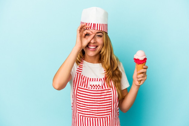 Caucasica gelatiera donna che tiene un gelato isolato su sfondo blu eccitato mantenendo il gesto ok sull'occhio.