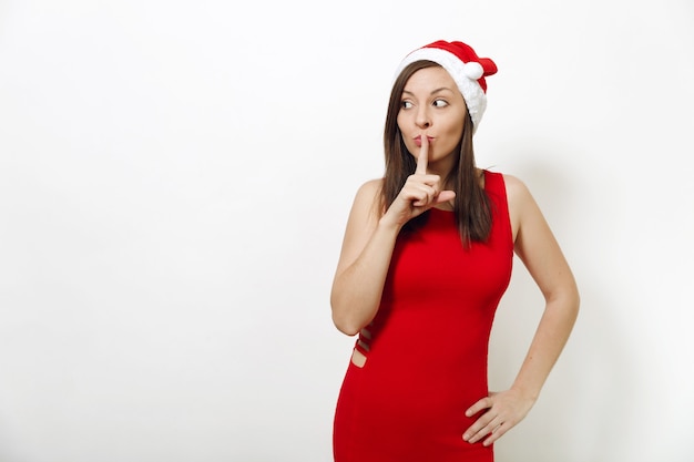 Caucasica giovane donna felice che indossa abito rosso e cappello di Natale tenendo il dito indice davanti alle labbra su sfondo bianco isolato. Ragazza di Santa che mostra gesto di silenzio. Concetto di vacanza di Capodanno 2018.