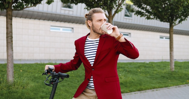 Caucasica giovane uomo bello in giacca rossa in piedi sulla strada in bicicletta o scooter elettrico e bere caffè. Ragazzo alla moda che sorseggia un drink per andare all'aperto. Bicicletta. Fermata del ciclista per riposare.