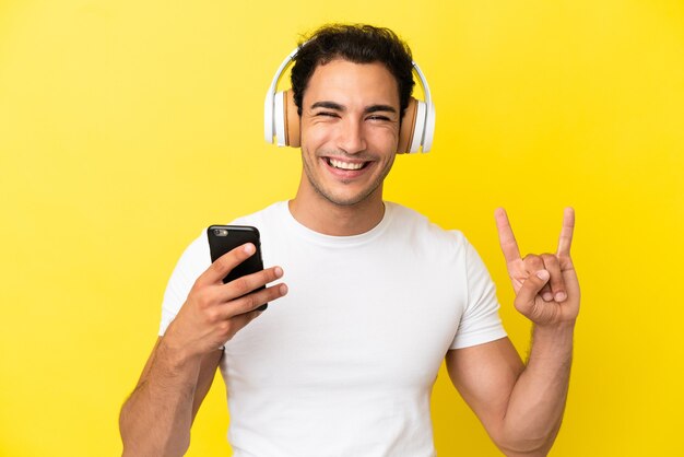 Caucasico bell'uomo su sfondo giallo isolato ascoltando musica con un cellulare che fa gesto rock