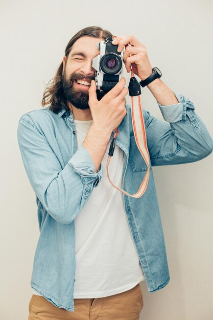 Caucasico bello positivo che scatta foto con la sua macchina fotografica mentre è in piedi isolato contro il muro bianco