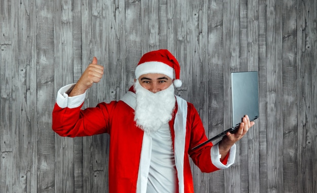 Caucasico giovane vestito da Babbo Natale con il pollice in alto e laptop in mano. Concetto di shopping online con 5 stelle