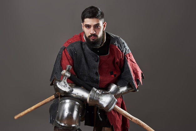 Cavaliere medievale su grigio. Ritratto di brutale guerriero faccia sporca con cotta di maglia armature rosse e nere e ascia da battaglia