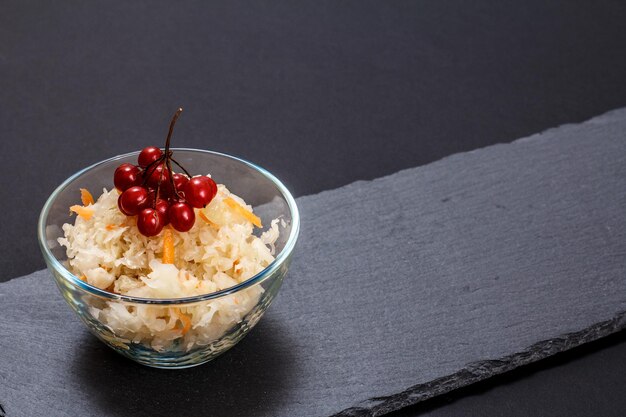 Cavolo fermentato fatto in casa con carota e grappolo di viburno in una ciotola di vetro sullo sfondo nero. Insalata vegana. Il piatto è ricco di vitamina U. Ottimo cibo per una buona salute.