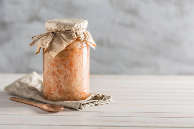 Cavolo fermentato in un barattolo di vetro con un cucchiaio di legno e un tovagliolo di lino su uno sfondo chiaro.