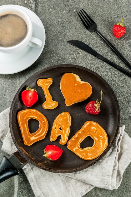 Celebrando la festa del papà. Prima colazione. L'idea per una ricca e deliziosa colazione: frittelle in forma di congratulazioni - Adoro papà. In una padella, una tazza di caffè e fragole. Vista dall'alto copyspace