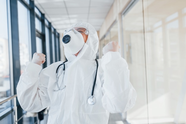 Celebrare la vittoria. Scienziato medico femminile in camice da laboratorio, occhiali difensivi e maschera in piedi al chiuso