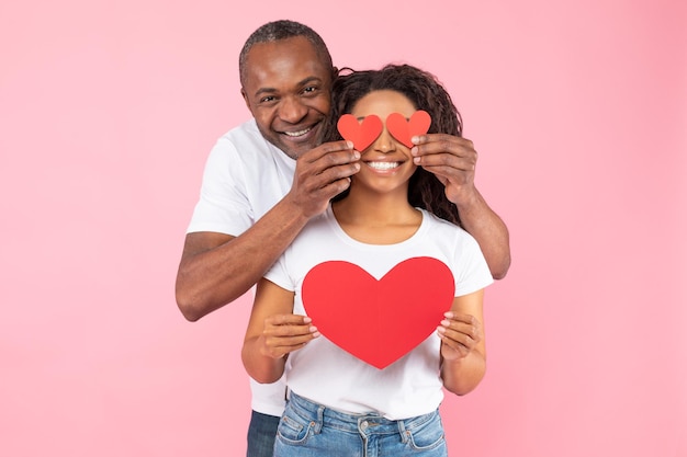 Celebrazione delle vacanze di San Valentino Amorevole marito nero di mezza età che copre gli occhi della moglie con cuori di carta