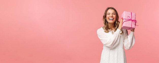 Celebrazione delle vacanze e concetto di donne Ritratto di felice ragazza bionda carismatica che agita la confezione regalo chiedendosi cosa c'è dentro come festeggiare il compleanno ricevere bday presenta sfondo rosa