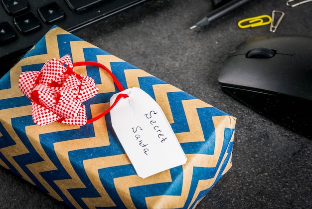 Celebrazione di Natale in ufficio, l'idea di condividere doni segreti di Babbo Natale. Tastiera, mouse, taccuino, penne, matite, regalo di Natale. Tavolo da ufficio nero, copyspace