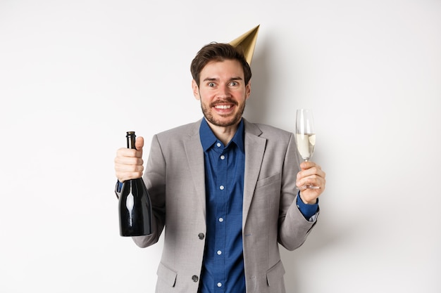 Celebrazione e concetto di vacanze. Ragazzo di compleanno bello eccitato in cappello da festa sorridente, bevendo champagne e divertendosi, sfondo bianco.