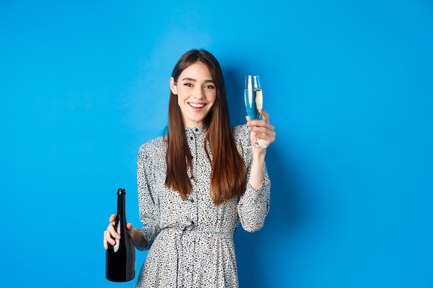 Celebrazione. Felice giovane donna in abito alzando un bicchiere di champagne, tenendo la bottiglia e sorridente, divertirsi alla festa, in piedi sul blu.